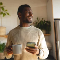 Young man in his kitchen, holding a mug of coffee and his phone.