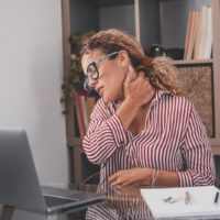 Woman sitting at a table with a laptop in front of her. She is holding her neck and has her eyes closed, as if in pain.