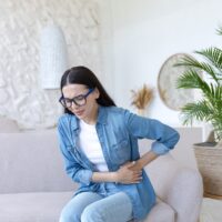 A young woman sits on a sofa at home. She appears to be in pain.